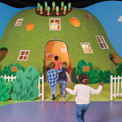 Toddler children are playing inside Rebecca Rabbit's house at PEPPA PIG World of Play Chicago. The colorful play area features a replica of Rebecca Rabbit's cozy home, complete with furniture and playful details. The children are interacting with the various elements of the house, including a kitchen and living area, while enjoying a hands-on, imaginative experience.